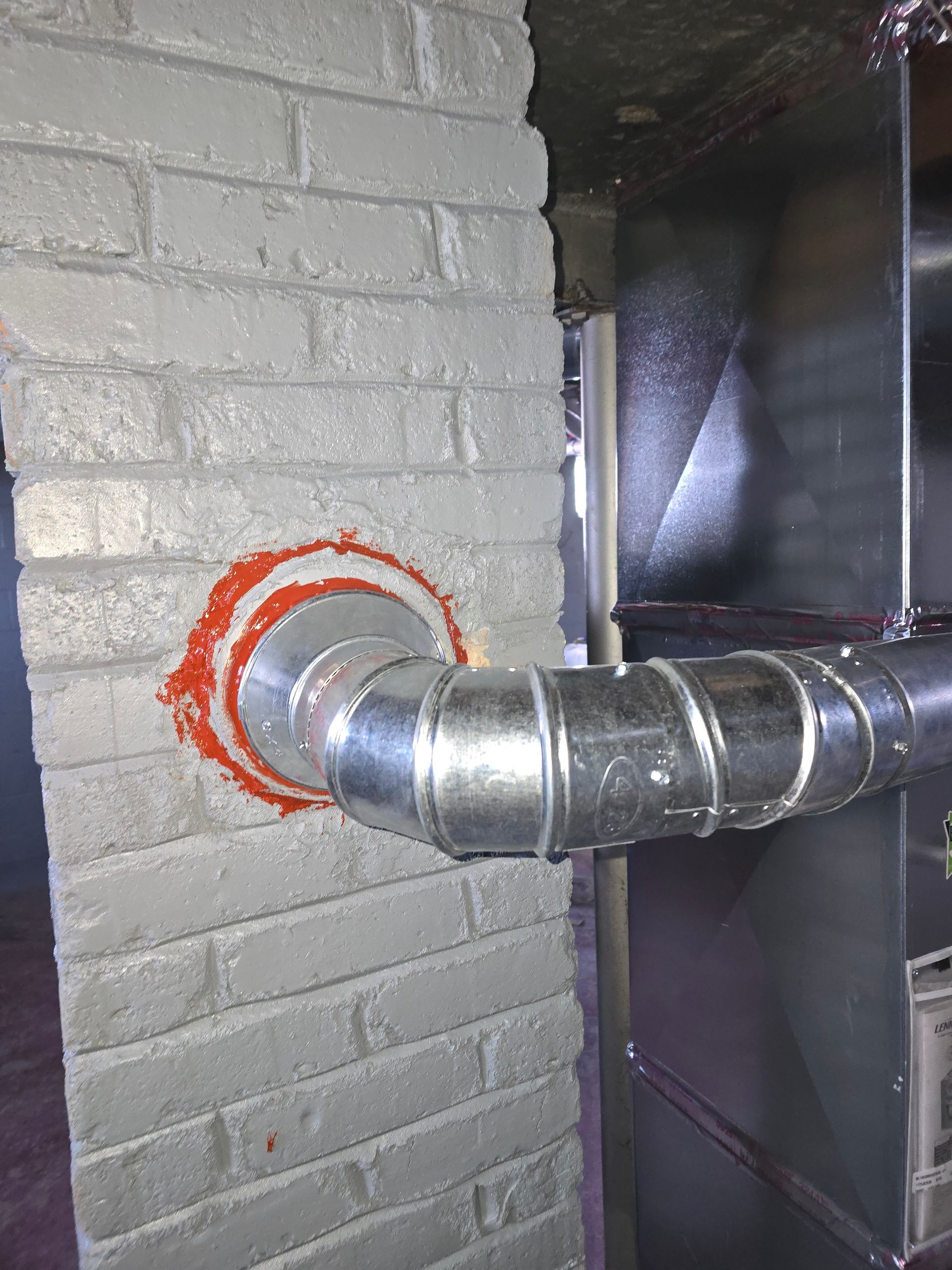 A metal ventilation pipe enters a brick wall, sealed with a thick ring of bright orange fire-rated sealant.