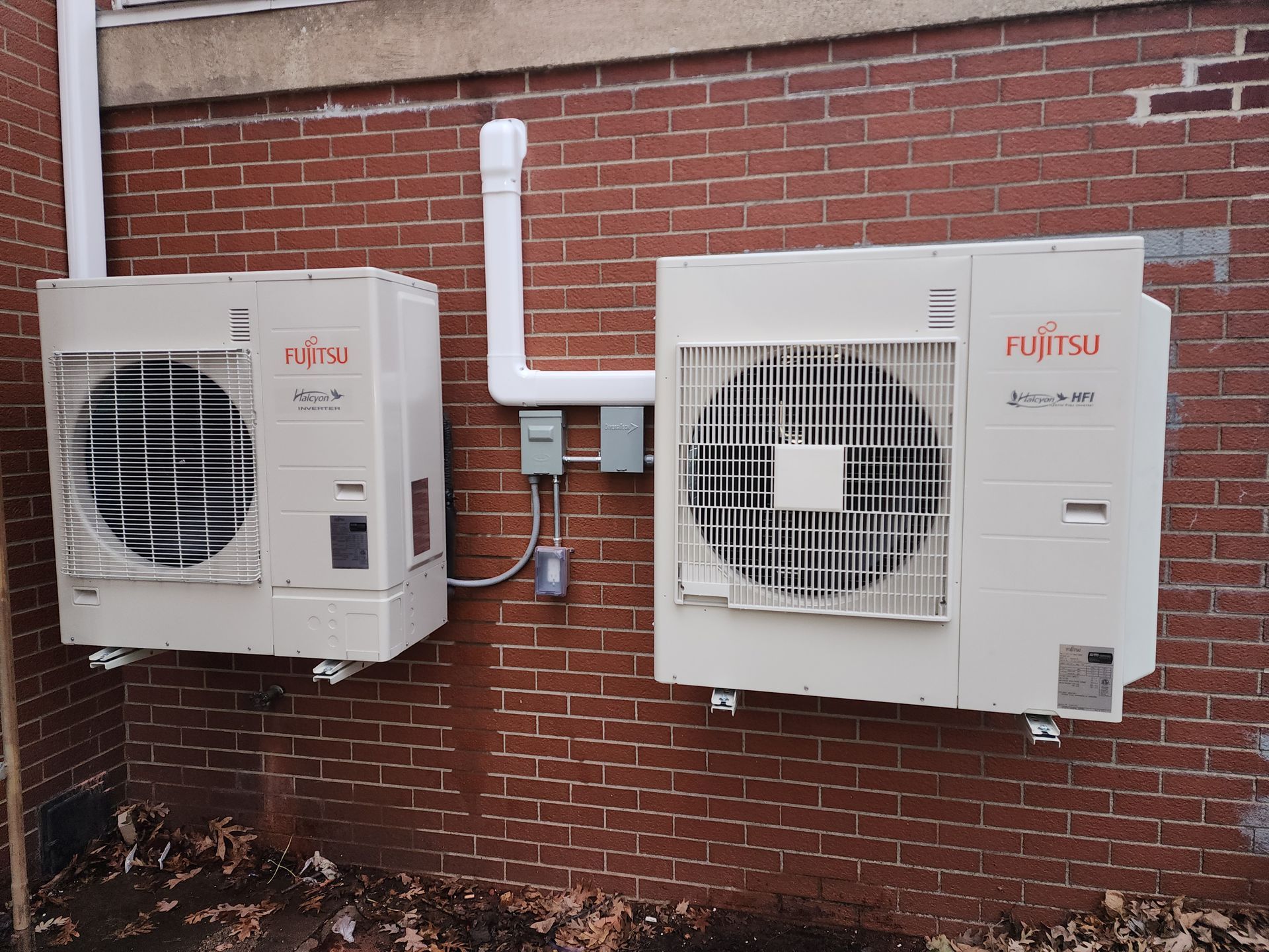 Two white Fujitsu outdoor HVAC units mounted on a red brick wall with associated electrical boxes and conduit.