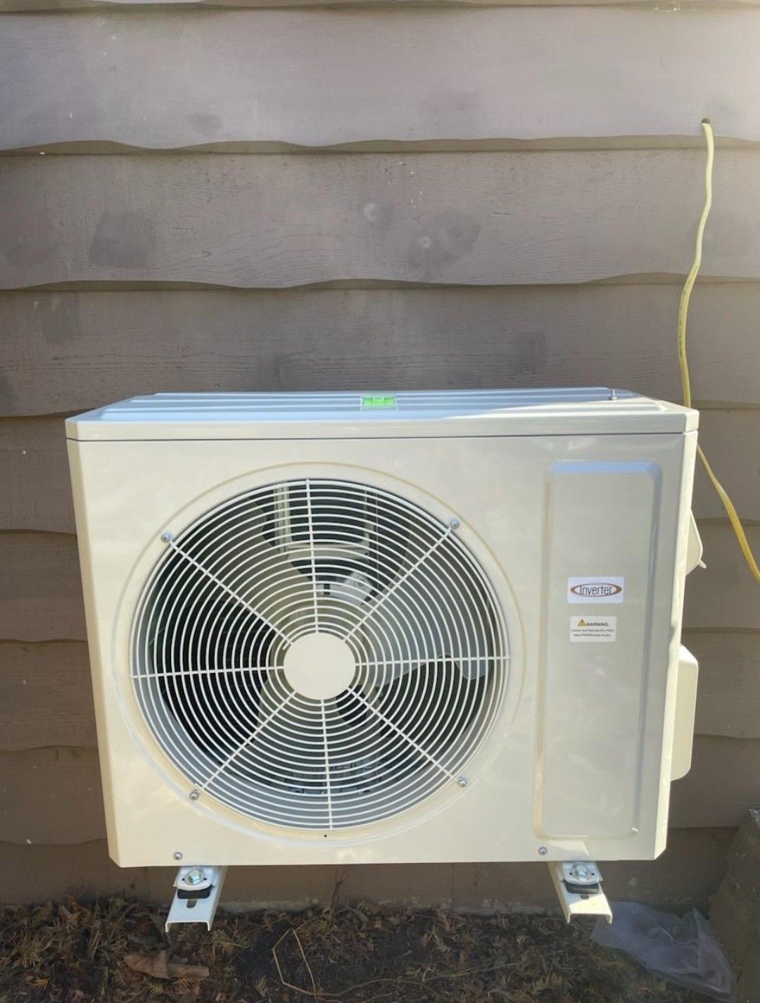 A beige outdoor AC condenser unit mounted on a wall with a visible yellow electrical wire running down the siding.