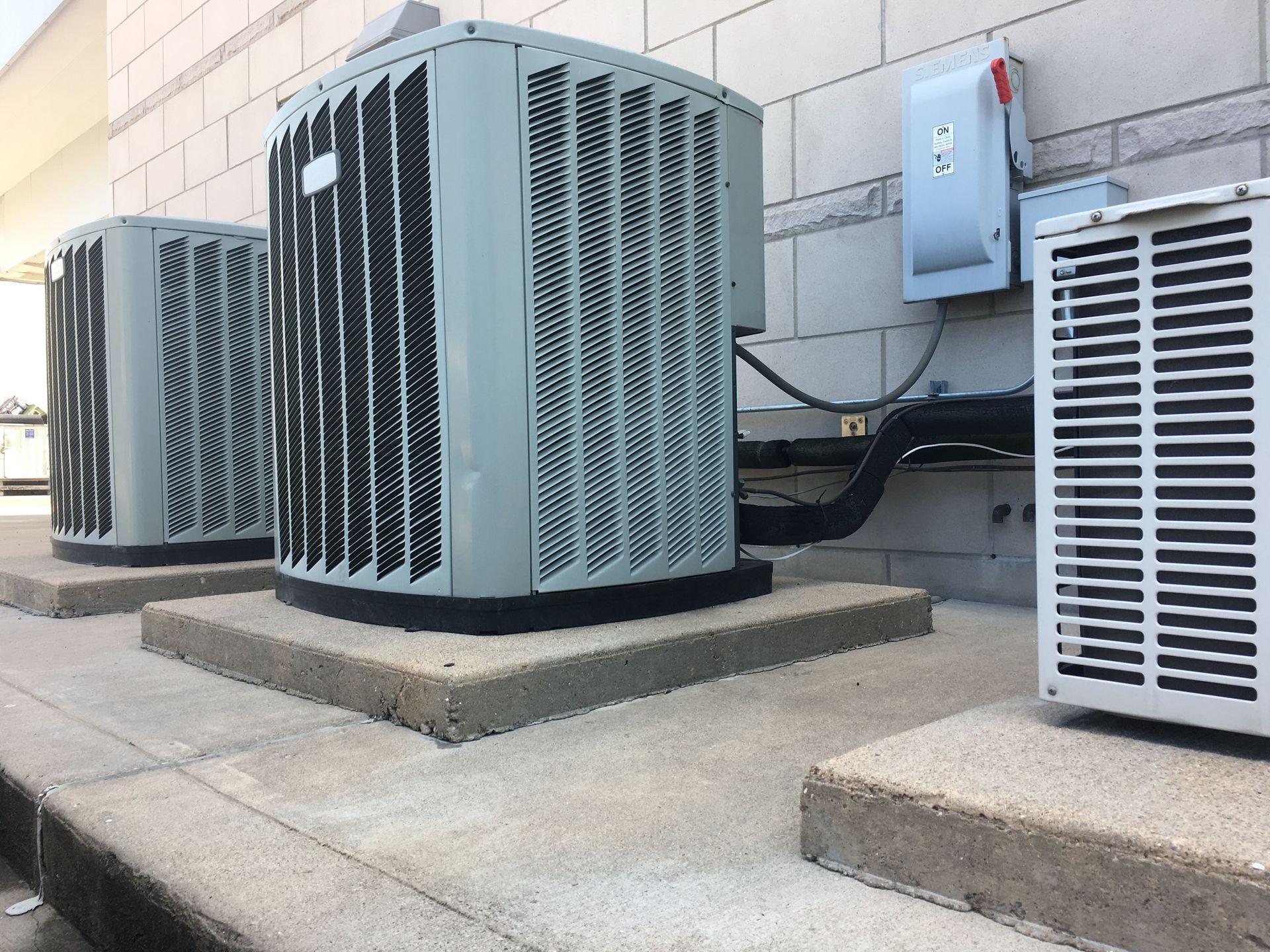Three grey outdoor HVAC condenser units sitting on concrete pads against a light-colored exterior wall.