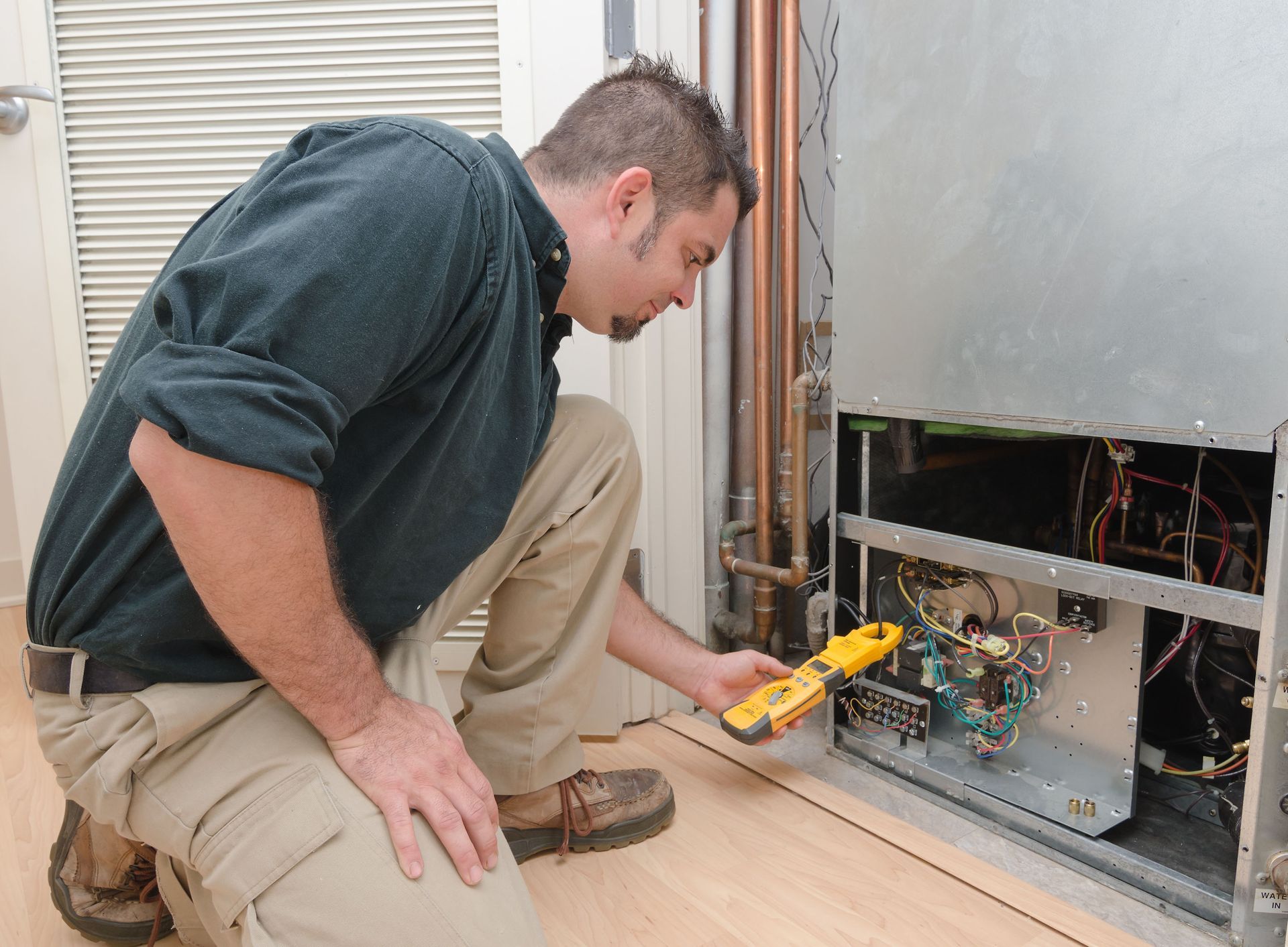 A technician in casual work clothes kneels, using a multimeter to test electrical components inside an HVAC unit.