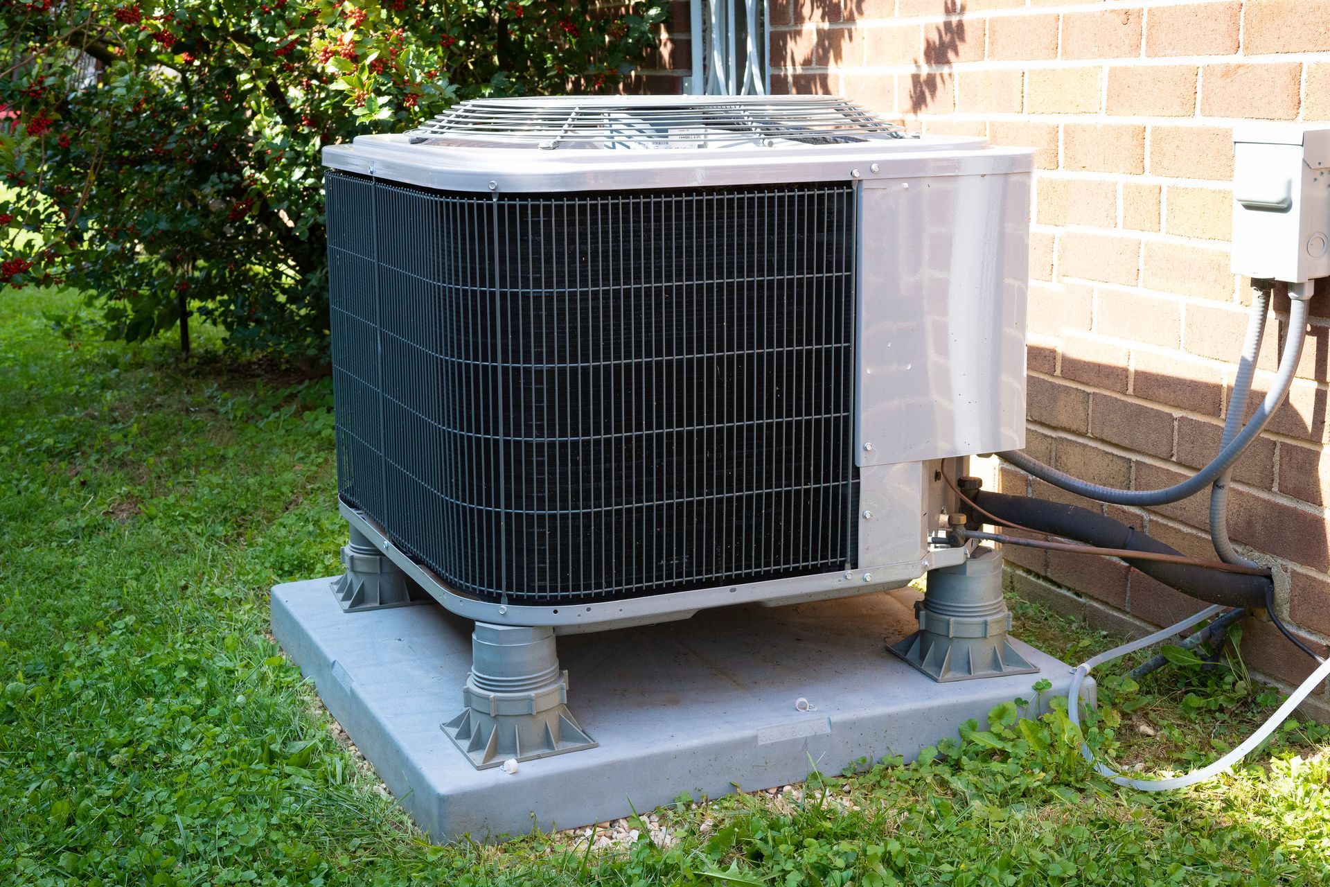 A grey residential air conditioning condenser unit sits on a concrete pad next to a brick house in a grassy yard.