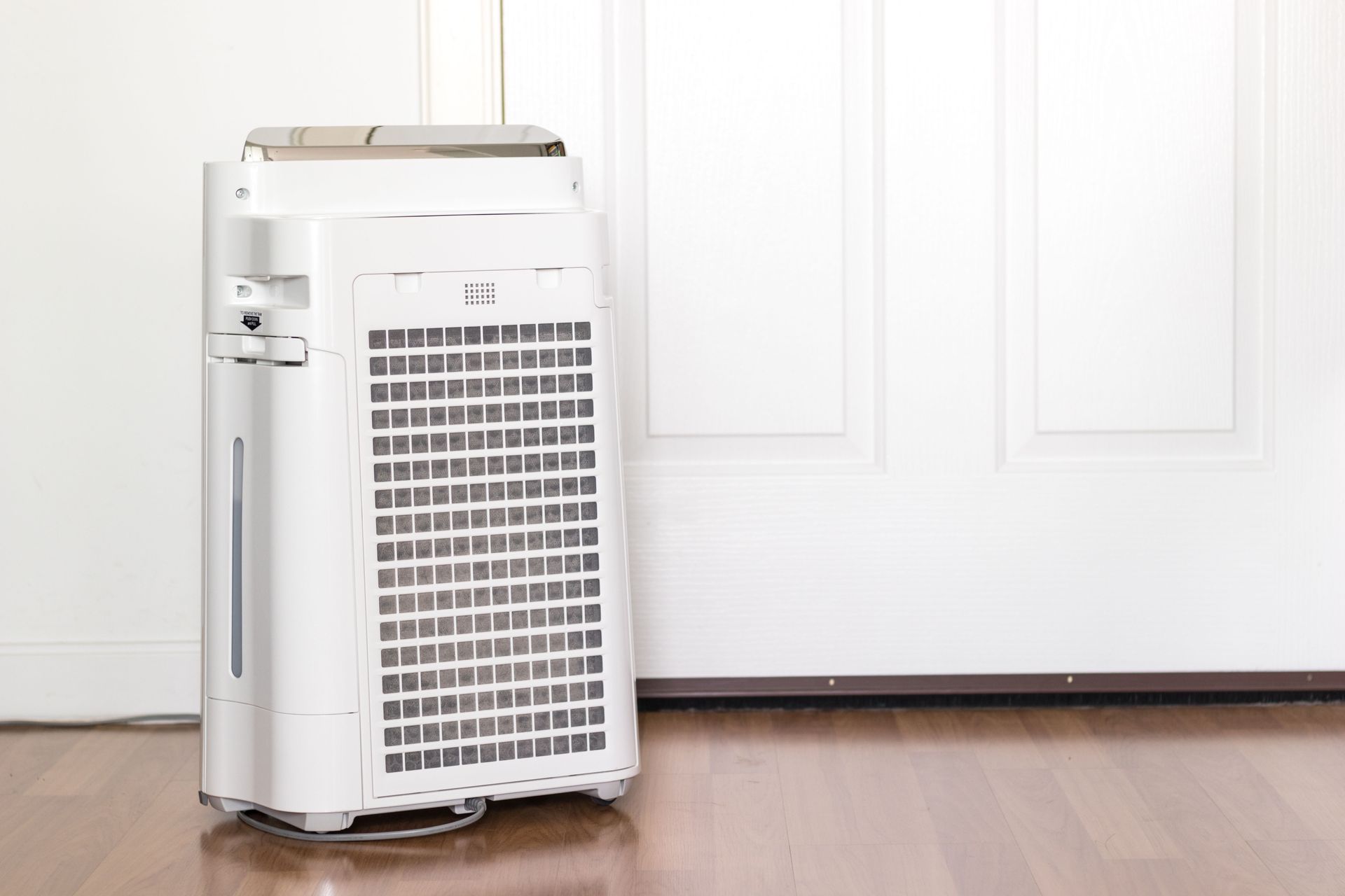 A white air purifier stands on a wooden floor in front of a white paneled door.