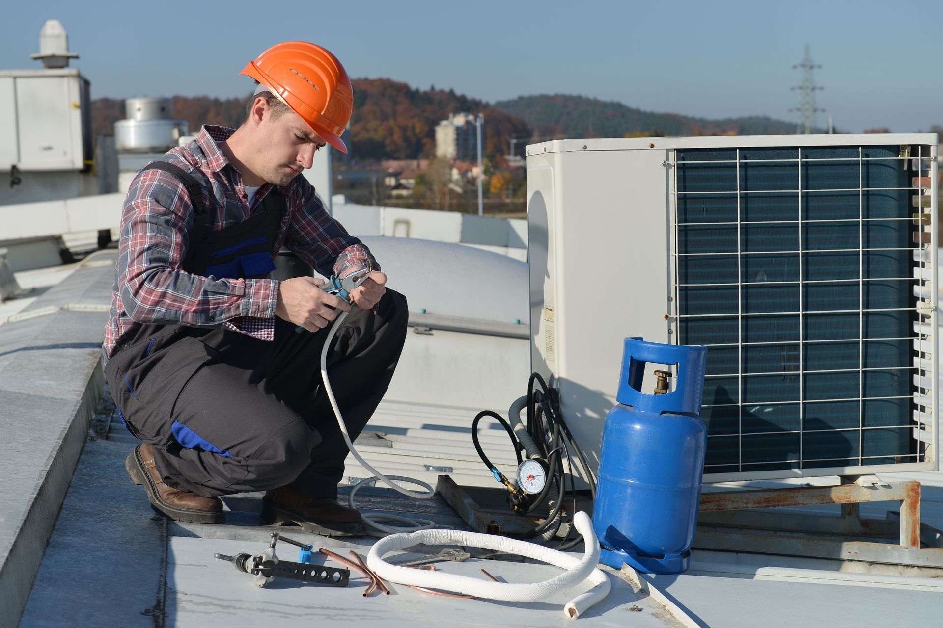 Technician in a hard hat kneeling on a rooftop, servicing an outdoor HVAC unit with gauges and a refrigerant cylinder.