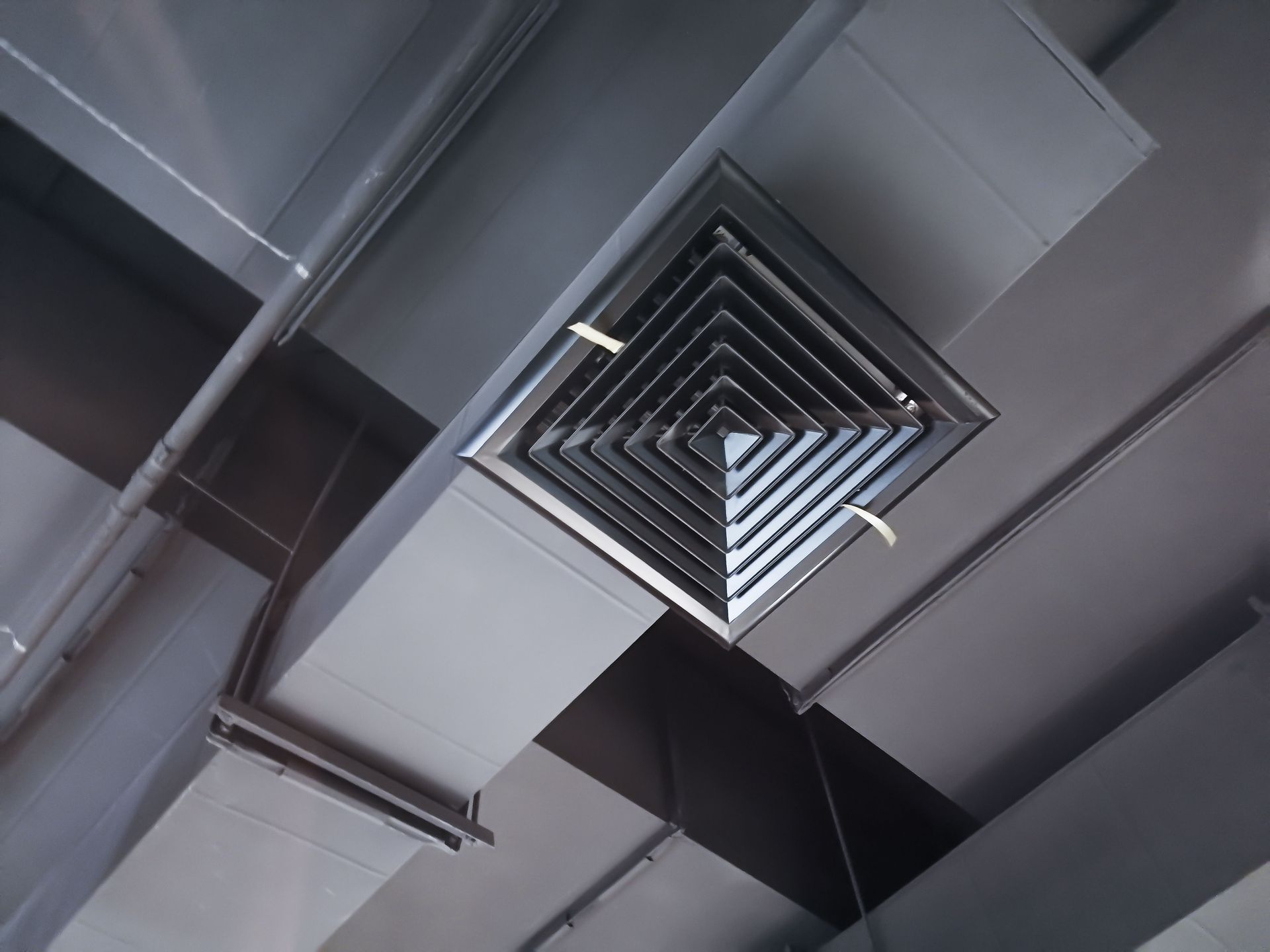 A square metal air conditioning vent is mounted to the ceiling, connected to a rectangular industrial ventilation duct.