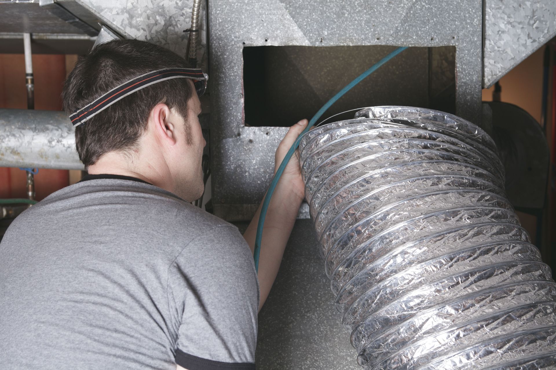 A worker in a gray shirt with a headlamp attaches a flexible metallic duct to a rectangular furnace opening.