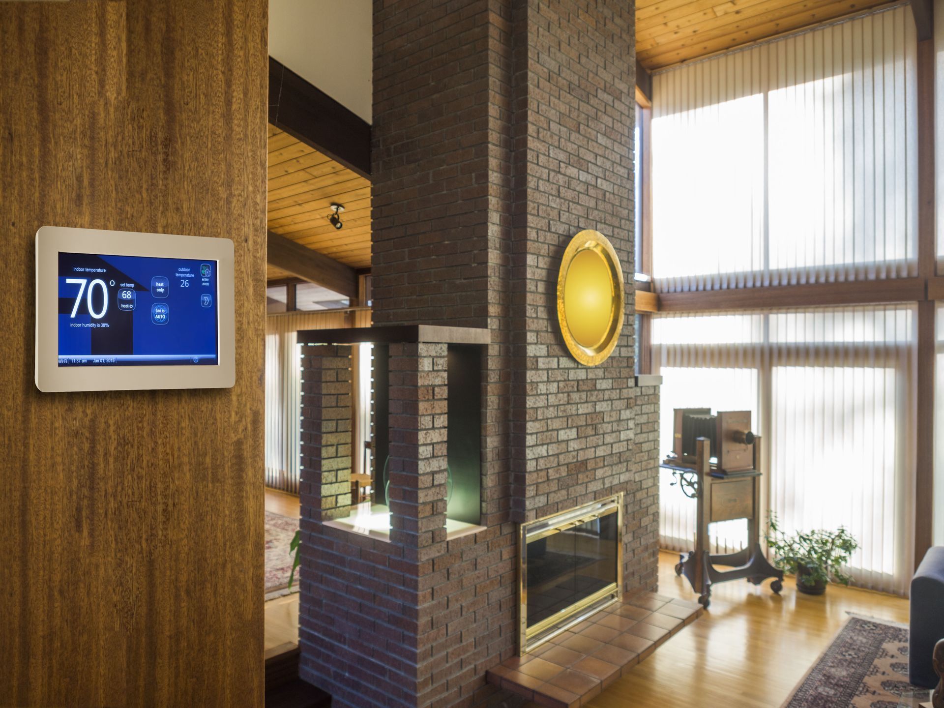 A smart thermostat mounted on a wood-paneled wall overlooking a living room with a brick fireplace and tall windows.