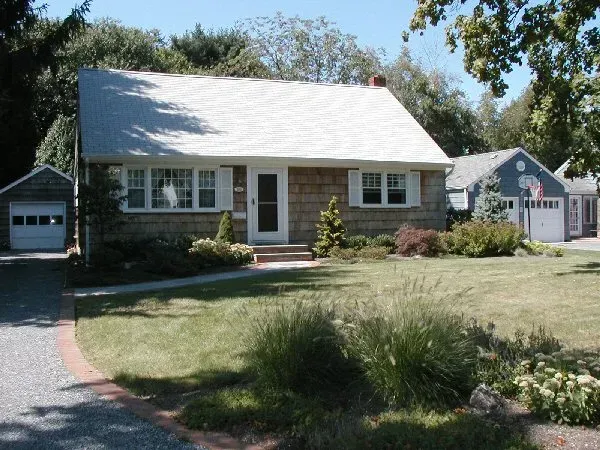 A single-story tan house with a white roof and trim, a small front lawn, and a gravel driveway with two detached garages.