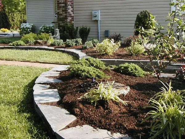 A curved flagstone border edges a mulched flower bed with small green shrubs and perennials in front of a residential home.