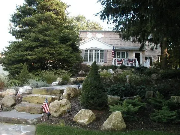 A two-story stone house with a landscaped tiered garden, large rocks, and a small American flag near the front steps.