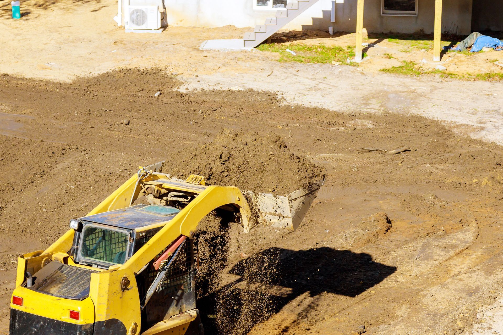 A yellow skid-steer loader scoops and moves piles of dirt at a construction site near a building foundation.