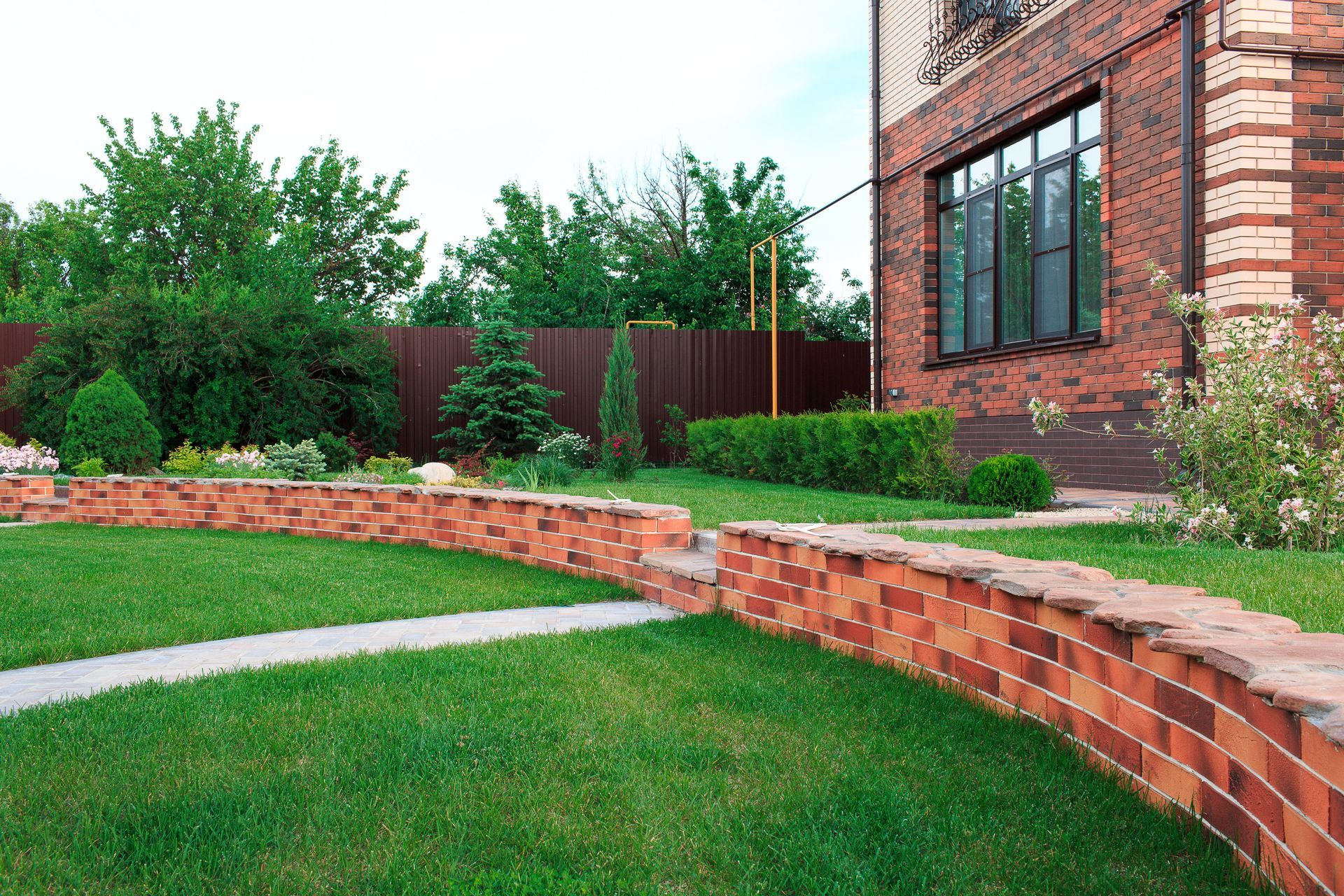 A curved brick retaining wall separates a green lawn from a landscaped garden next to a brick house.