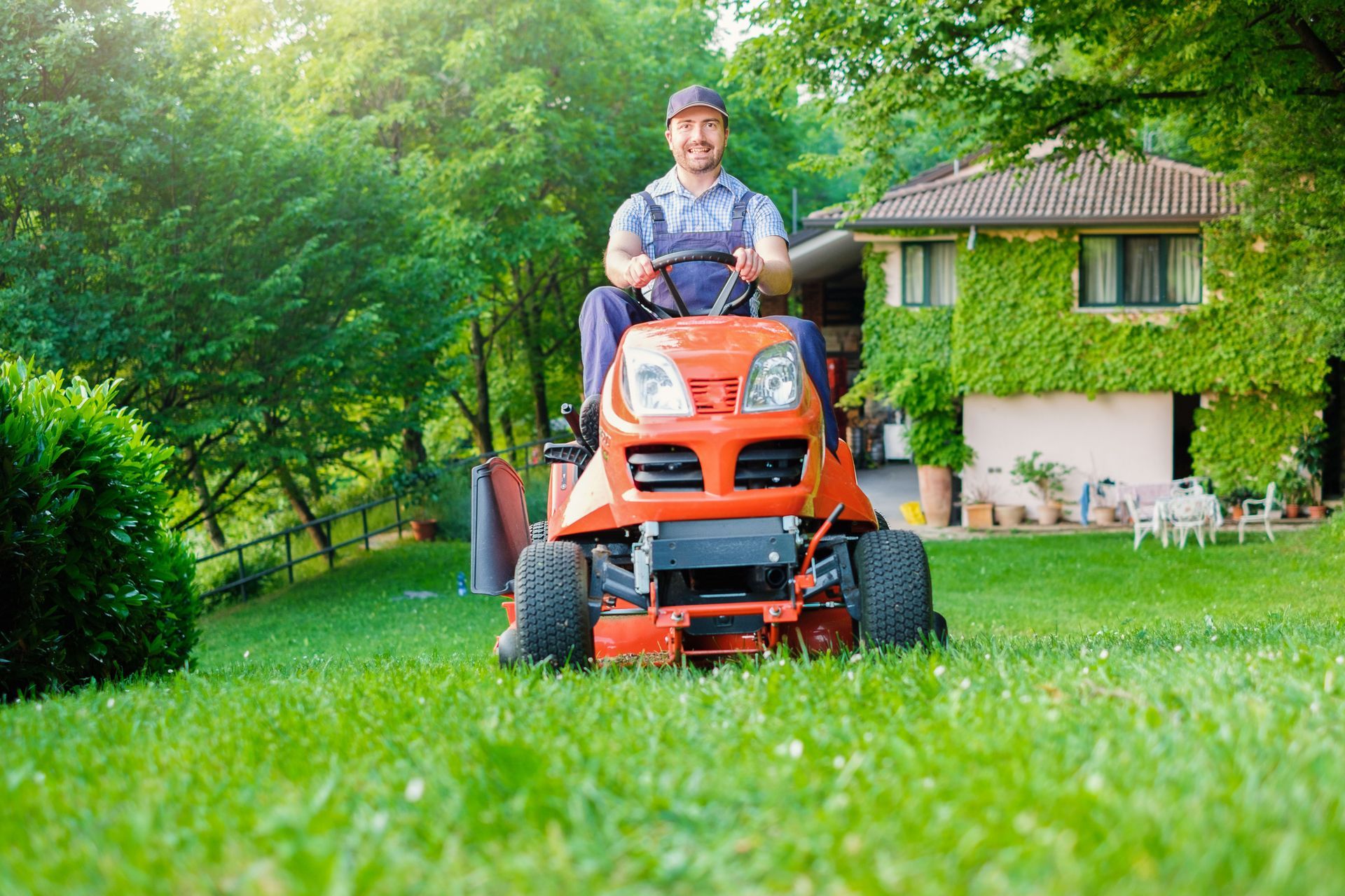 A person smiles while driving an orange riding lawn mower on a green grassy hill in front of a house.