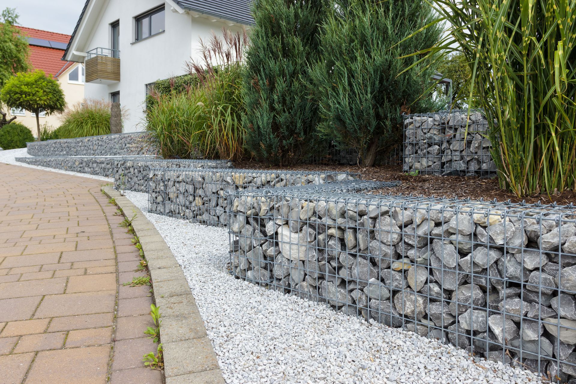 A modern home with a tiered landscape featuring stone-filled wire gabion retaining walls, white gravel, and shrubs.