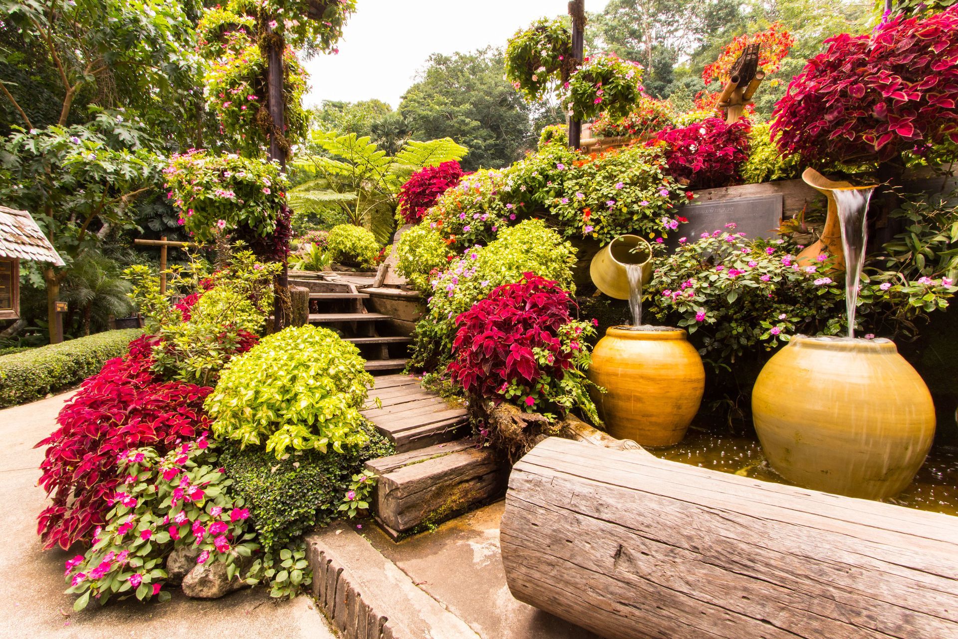 A tiered garden features vibrant red and lime green foliage, wooden steps, and two large ceramic pots with flowing water.