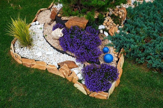 A garden bed features purple flowers, white gravel, blue spheres, and green conifers, all framed by stacked stone edges.