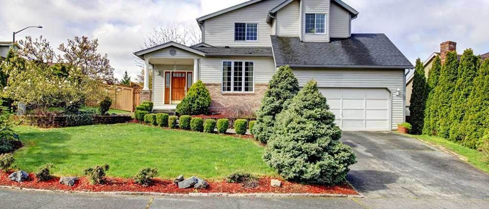 A two-story suburban house with a gray roof, white siding, a front lawn, a paved driveway, and evergreen landscaping.