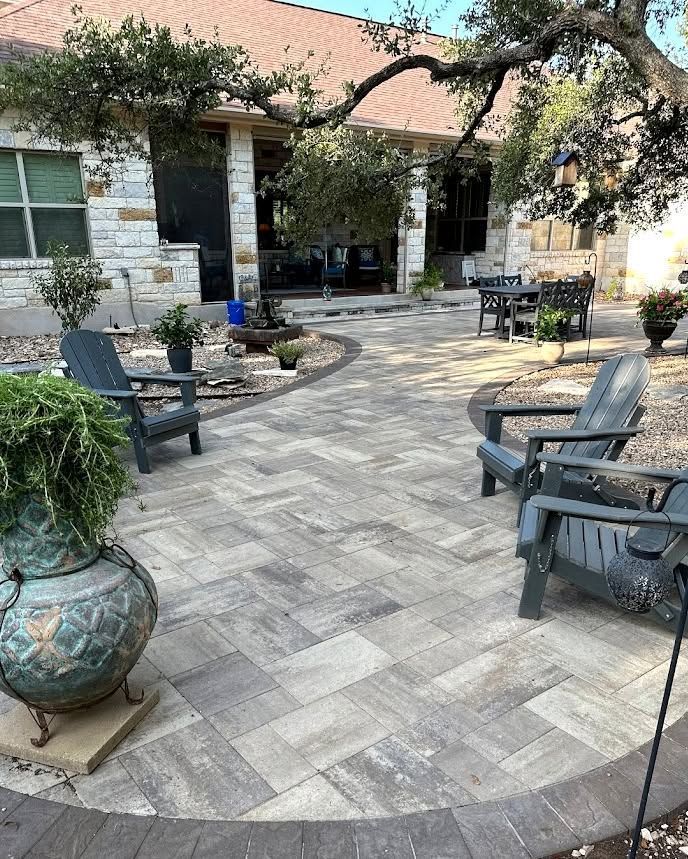 Patio with stone pavers, black chairs, potted plants, and a brick house entrance under trees.