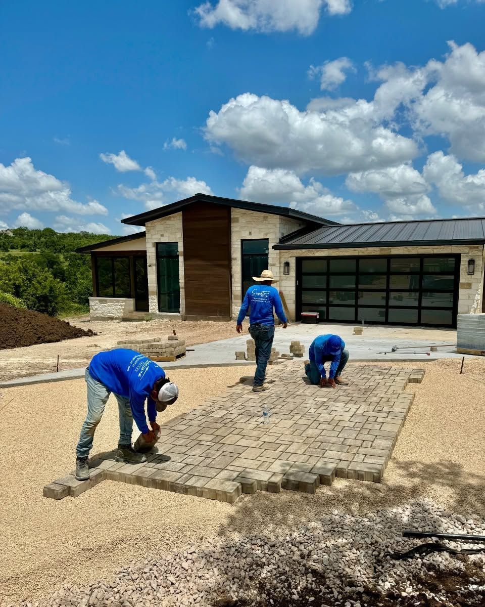 Workers laying pavers in front of a modern house under a bright blue sky.
