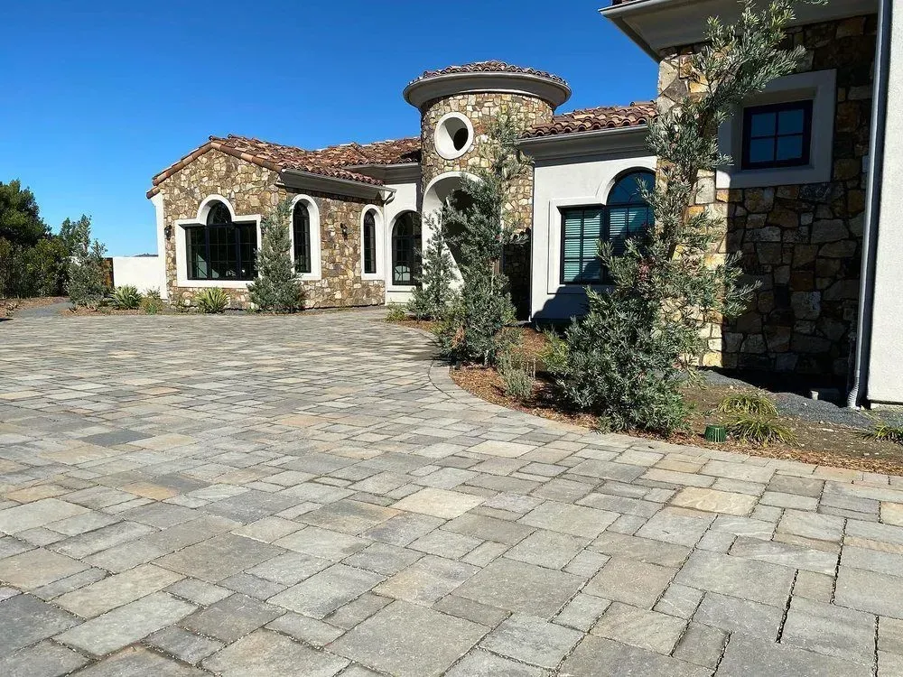 Stone Mediterranean-style house with arched windows and a wide paved driveway under a clear blue sky.