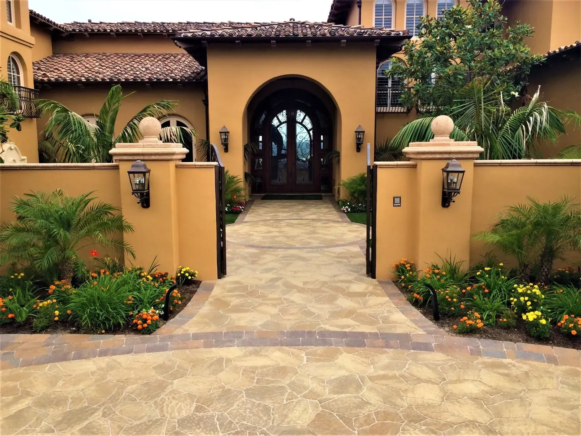 Mediterranean-style house entrance with arched doorway, stone path, and landscaped flower beds.