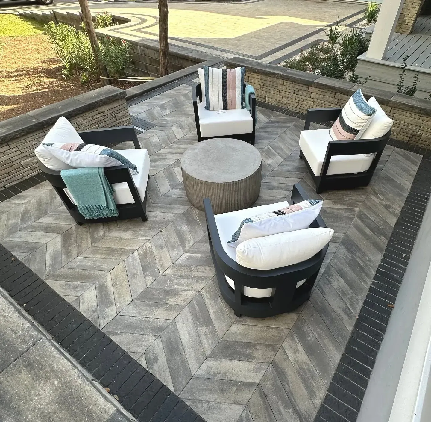 Outdoor patio seating area with four cushioned chairs around a round stone table on a herringbone paver patio.