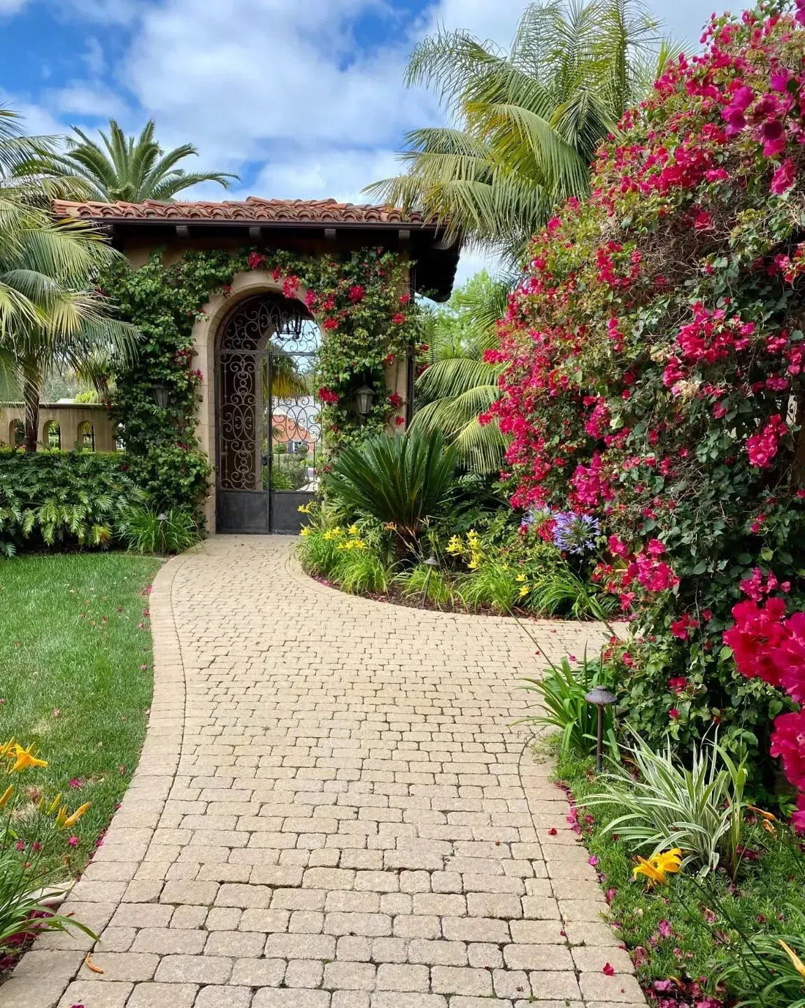 Curved garden path leading to a stone archway, with lush greenery and bright pink bougainvillea on the right.