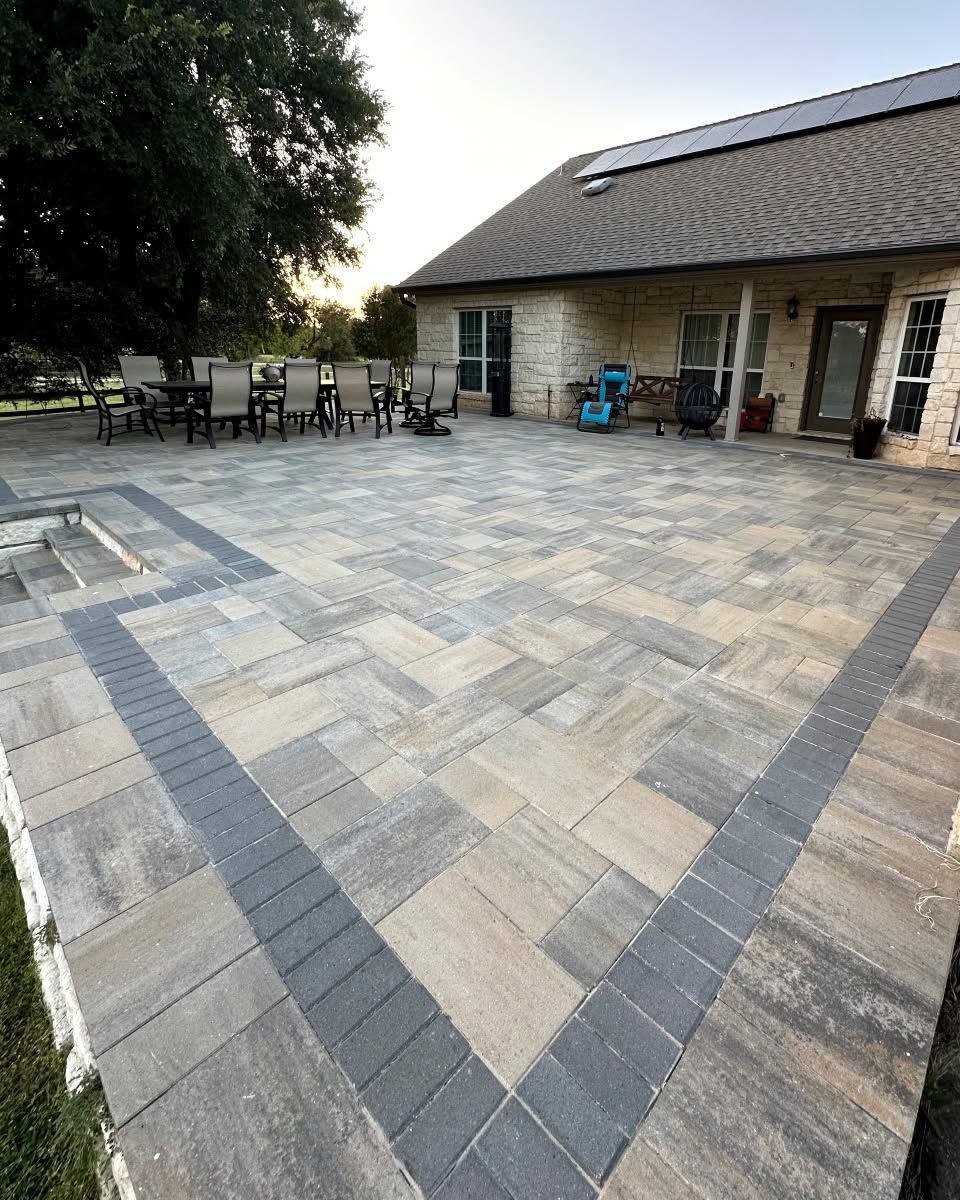 Stone patio with dark border tiles leading to a brick house and outdoor seating under trees.