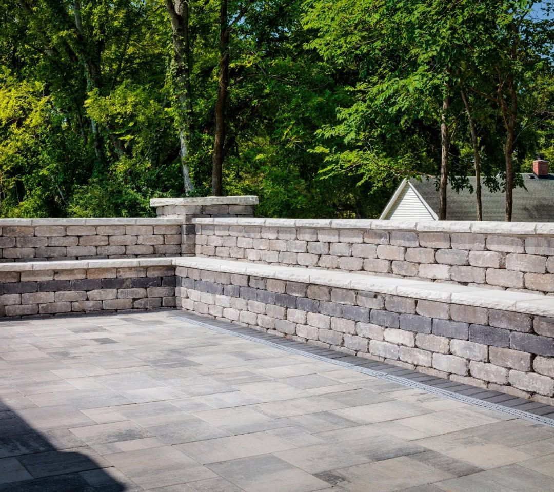 Stone patio with low brick retaining walls beside green trees in a park-like setting