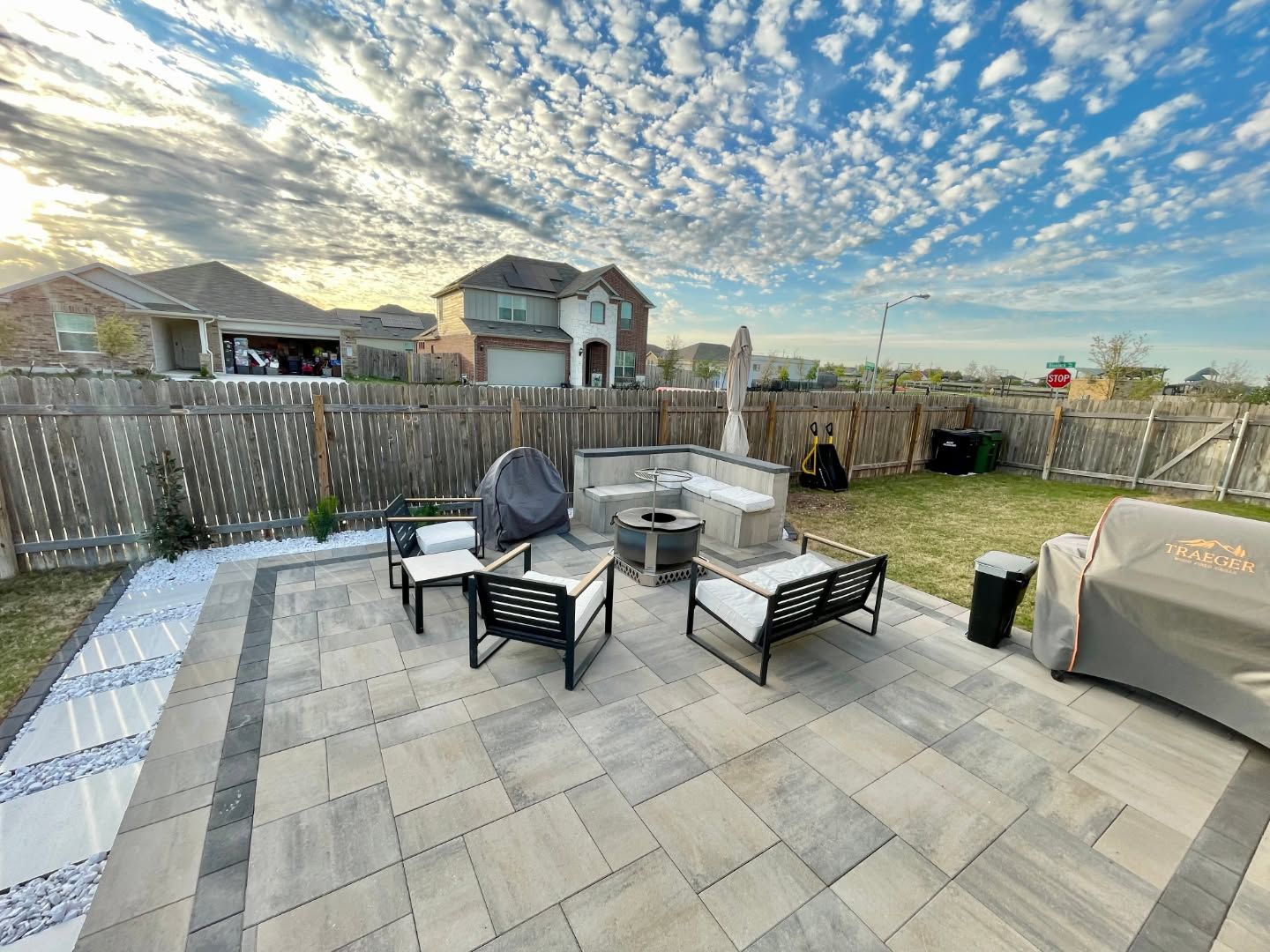 Patio with outdoor seating, grill, and fire pit under a cloudy sky, surrounded by fenced backyards.