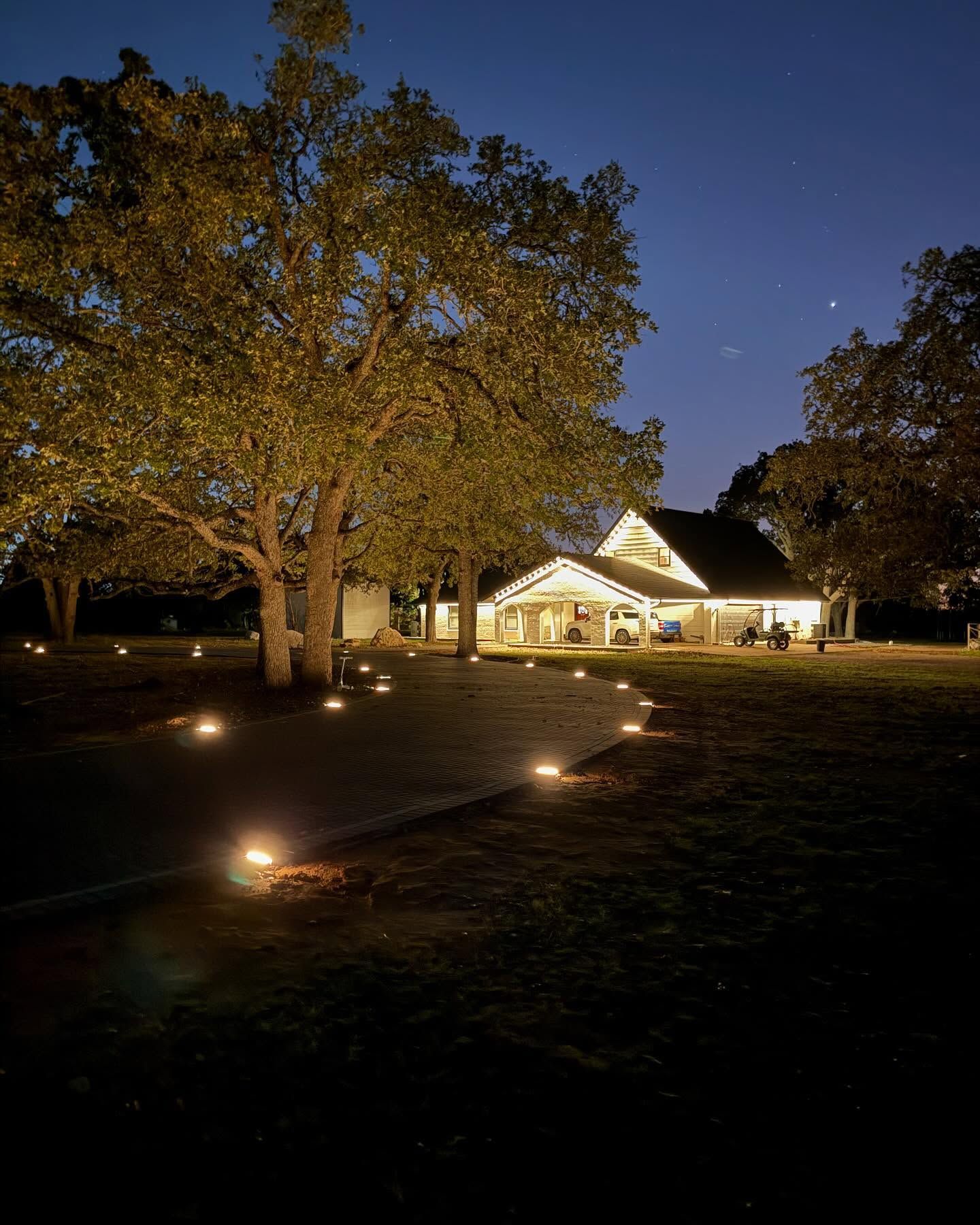 Nighttime park path with ground lights, trees, and a lit pavilion under a dark blue sky