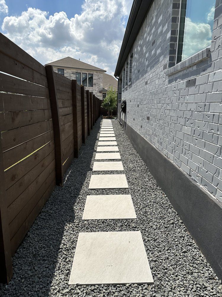 Narrow gravel side yard with stepping stones between a wooden fence and a brick house under a blue sky