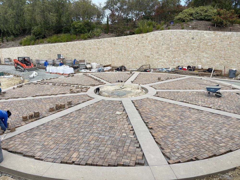 Circular brick-paved plaza under construction with radial concrete paths and stone retaining wall.