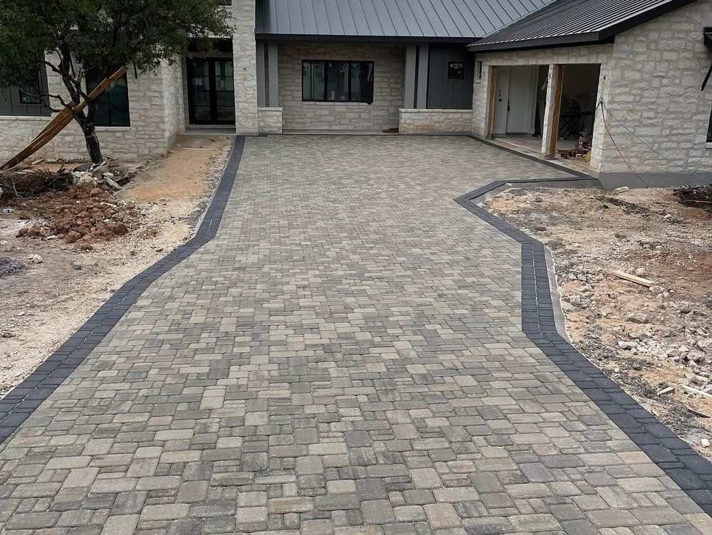 Stone paver driveway leading to a house entrance, bordered by dark concrete edging and unfinished landscaping