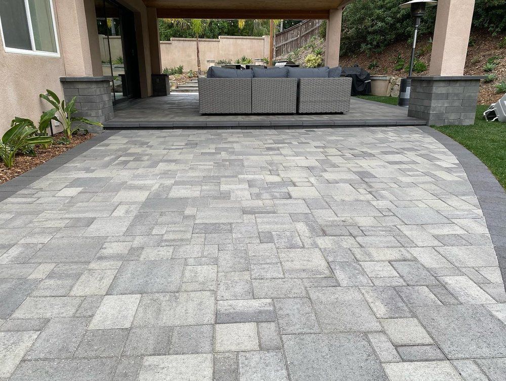 Gray stone-paver driveway leading to a covered patio with outdoor seating and greenery on the sides
