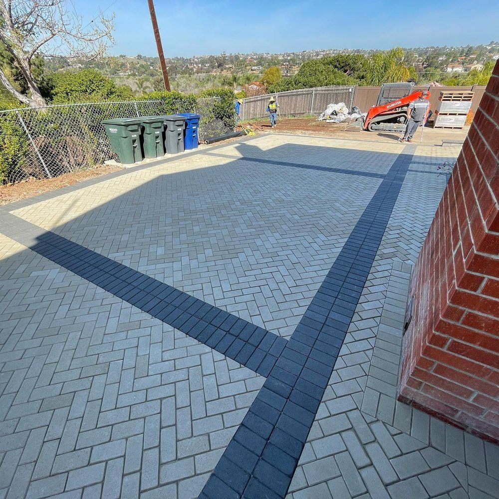 Backyard patio with light pavers, dark border, brick wall, fence, bins, and red chairs in the distance