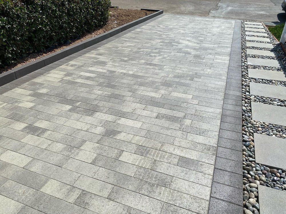 Paved driveway with light gray pavers and a gravel border beside trimmed hedges