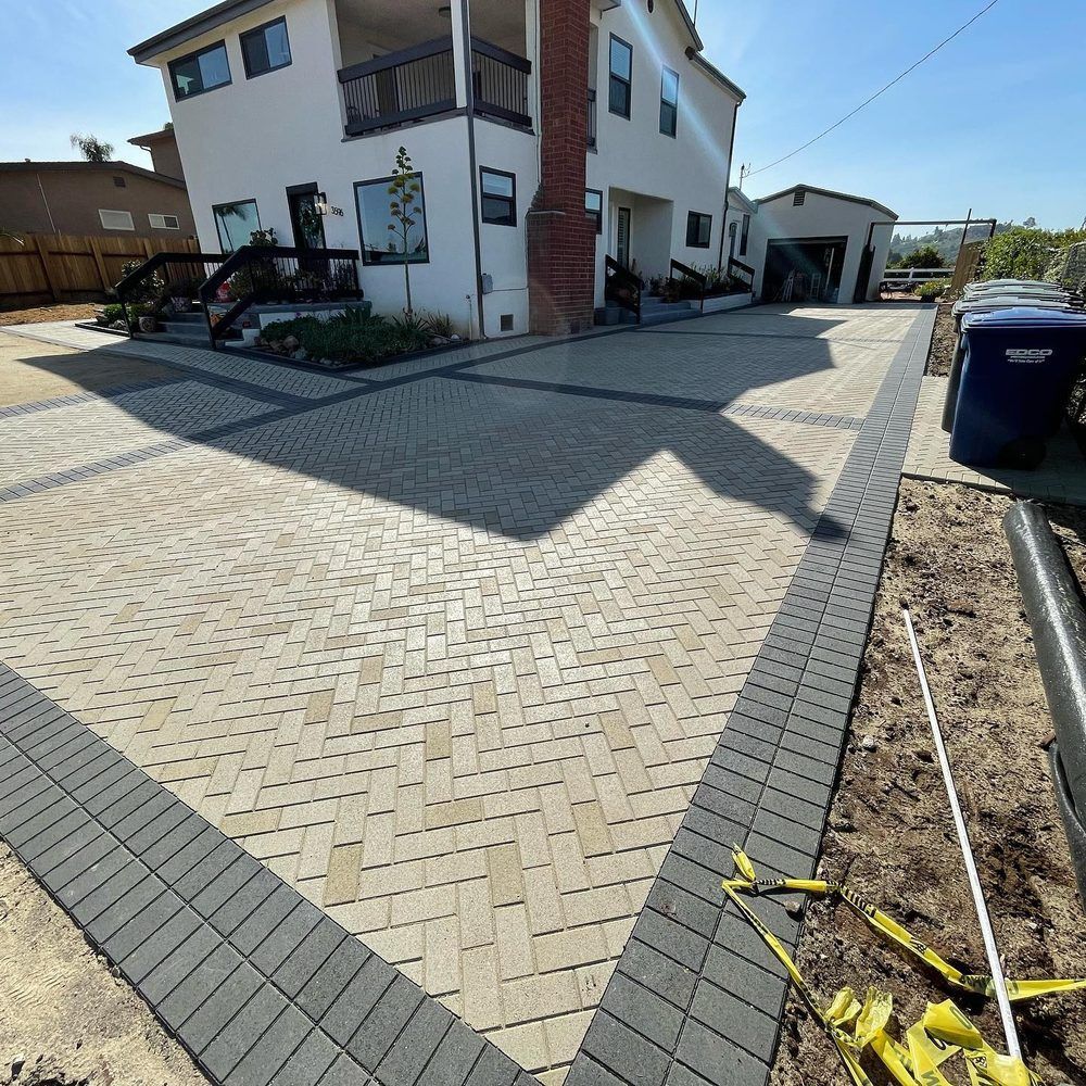 Brick-paved driveway beside a white house with a garage and trash bins on a sunny day