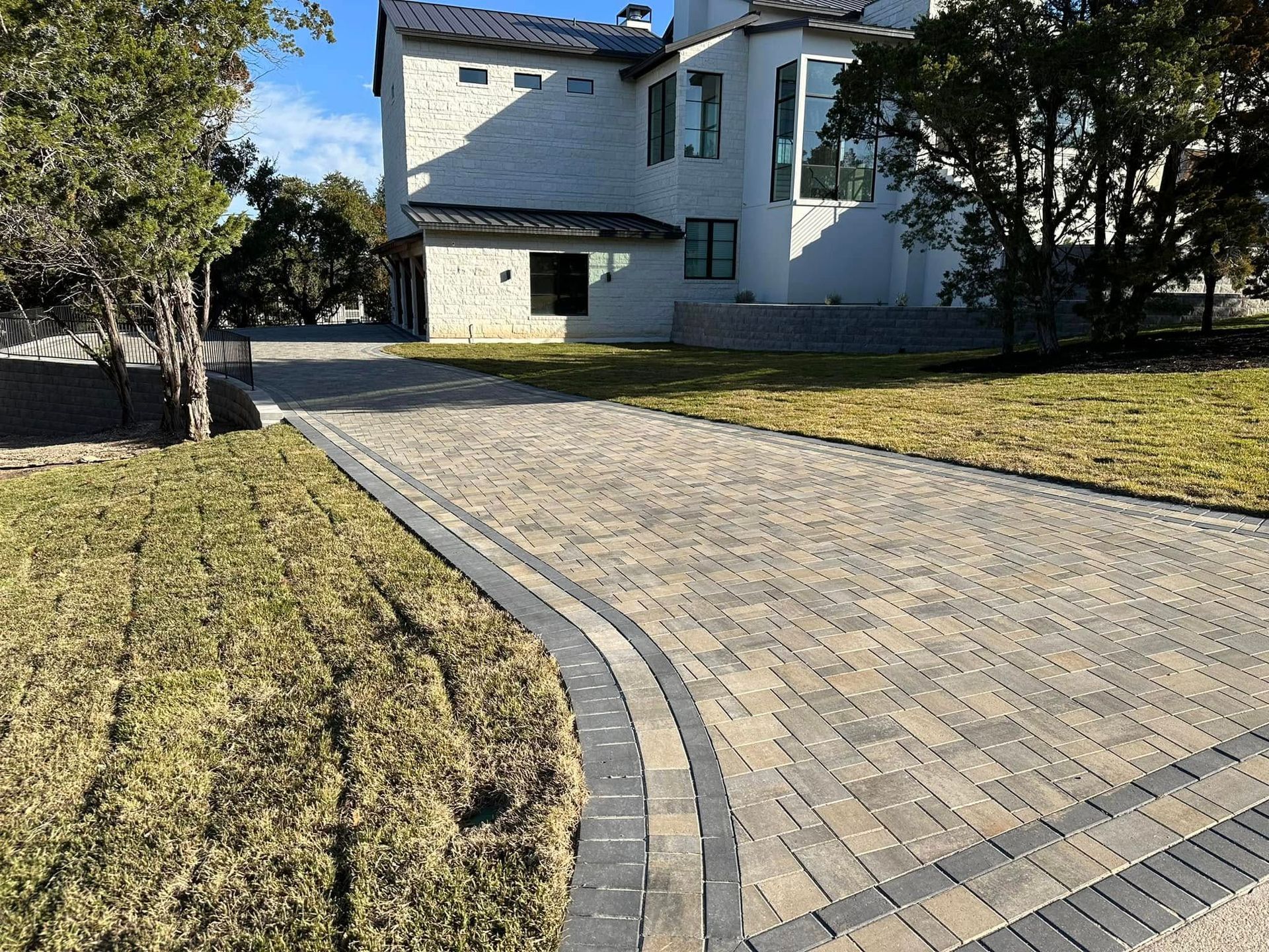 Brick driveway leading to a white house with trees and lawn on a sunny day