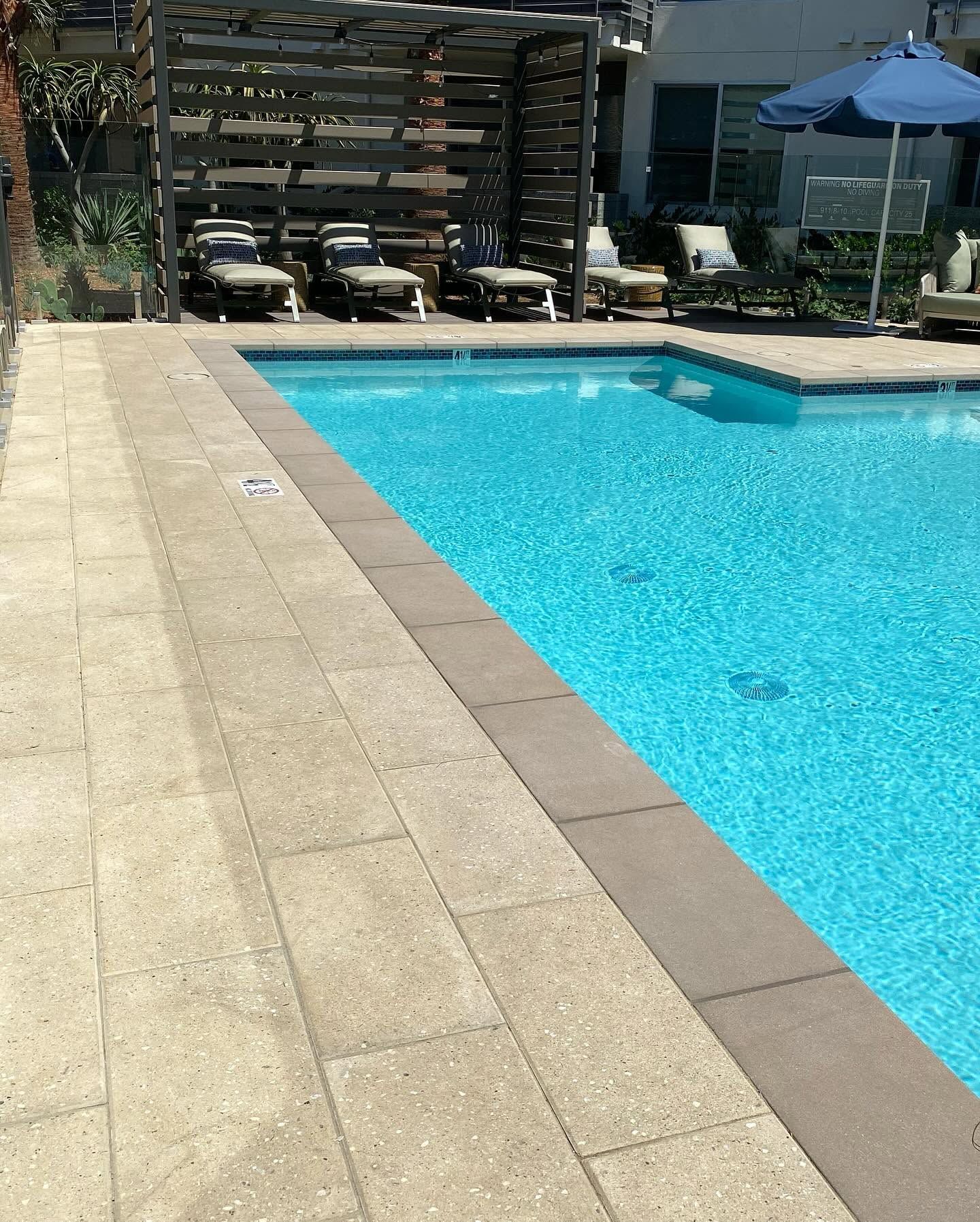 Outdoor swimming pool with blue water beside a beige tiled deck and lounge chairs in the background