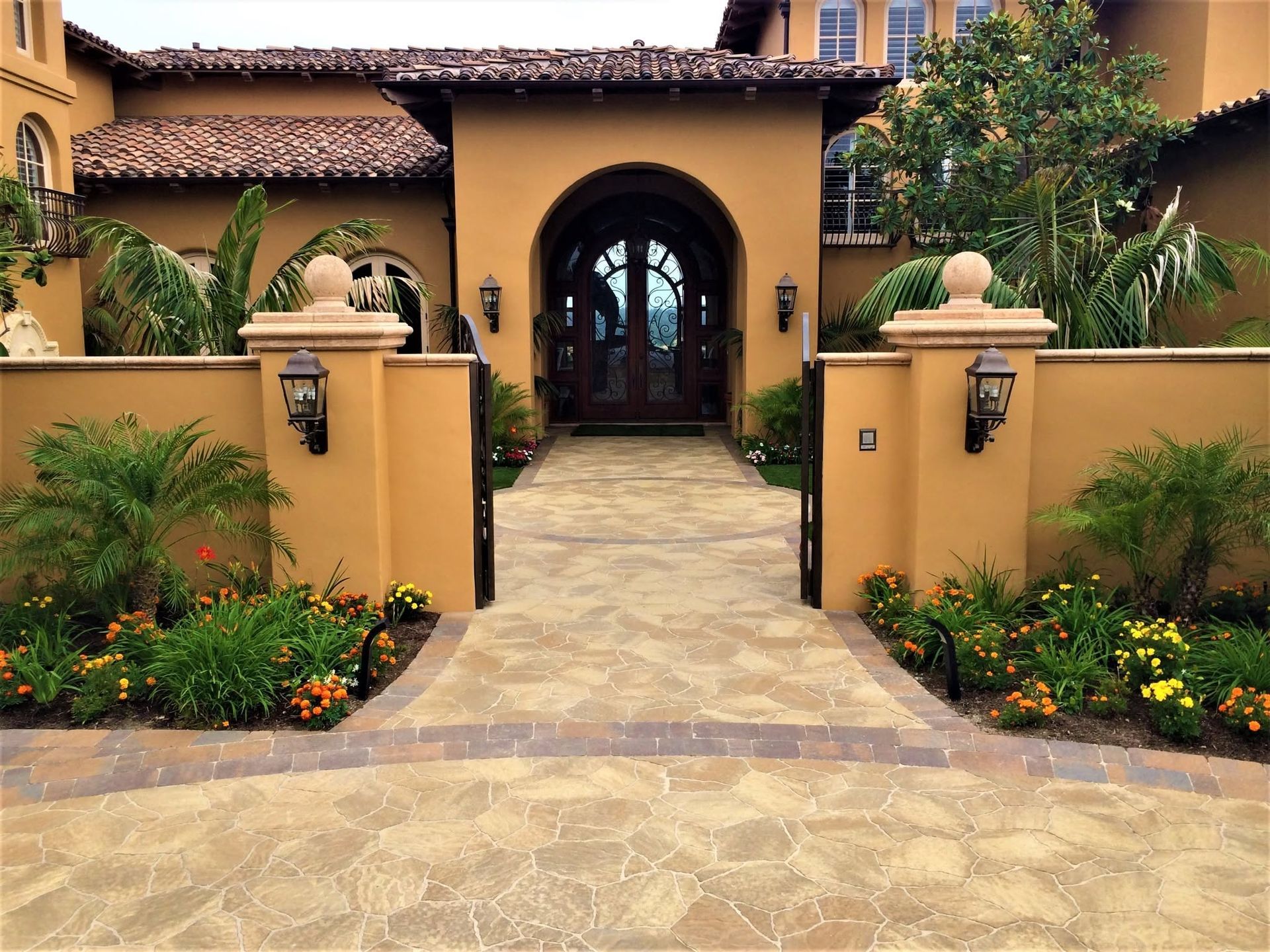 Stone walkway to a tan stucco home with an arched doorway, flanked by garden walls and lush landscaping