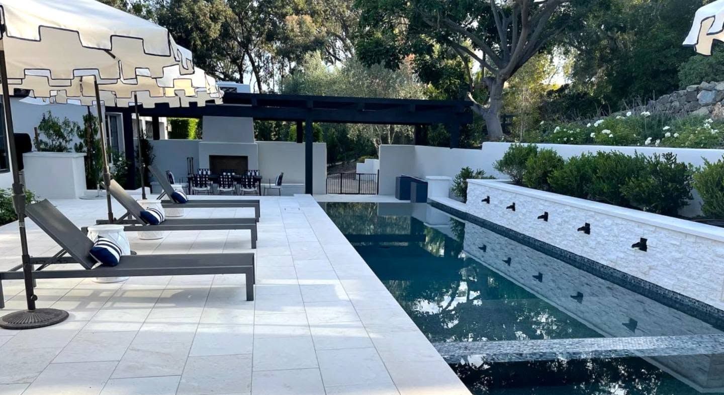 Modern backyard with a long rectangular pool, white lounge chairs, and a pergola seating area with trees beyond.