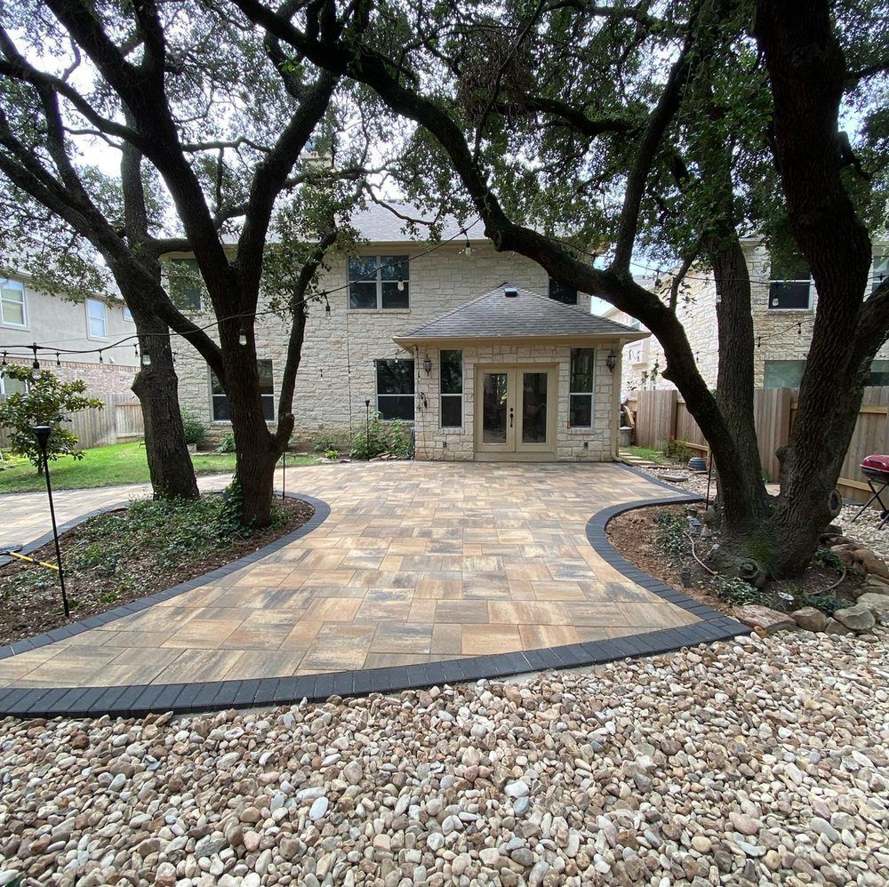 Stone patio walkway leading to a two-story house framed by large trees and landscaped gravel beds