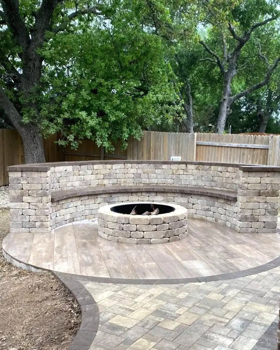Outdoor stone fire pit with curved bench seating in a fenced backyard under trees