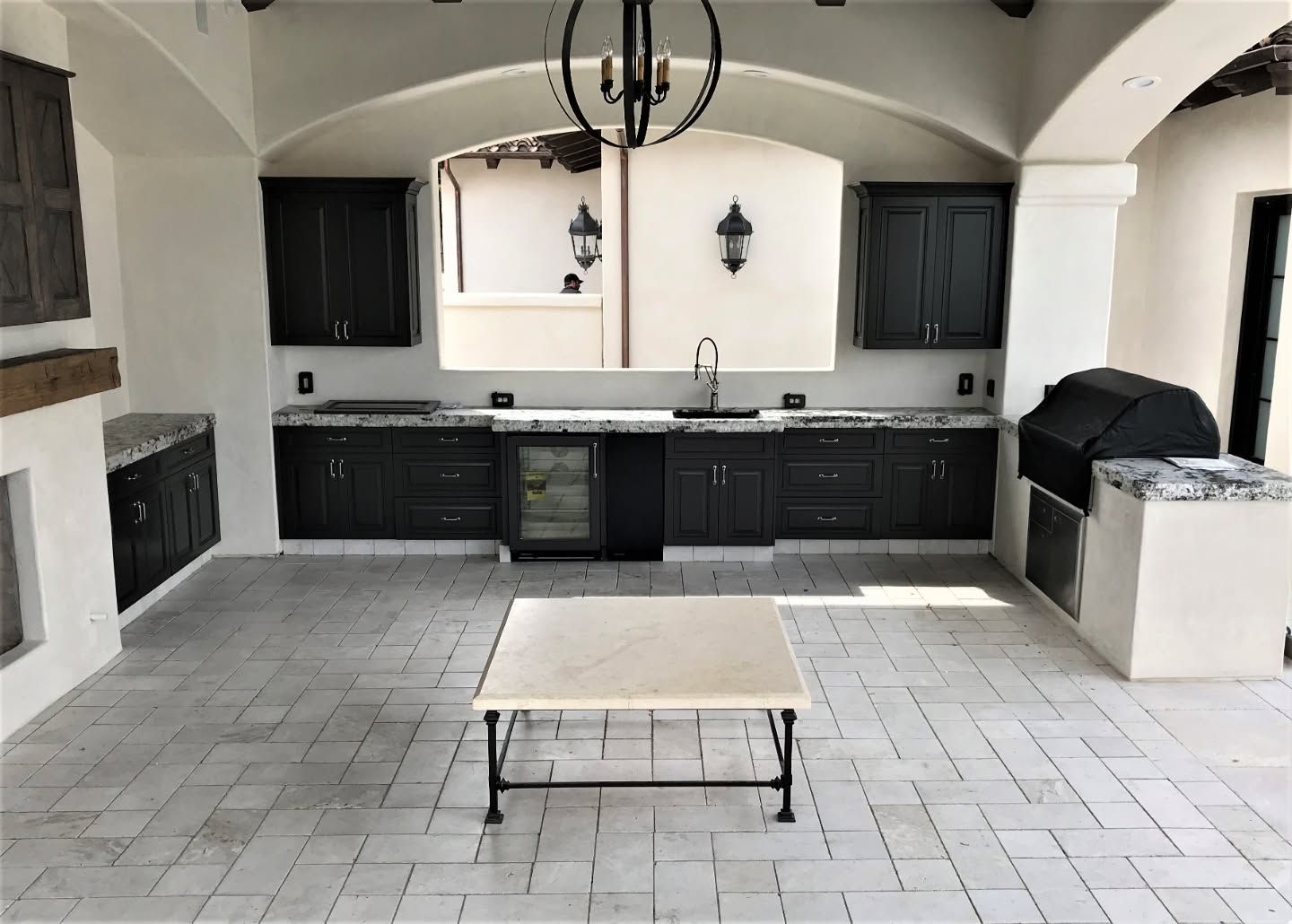 Outdoor patio kitchen with dark cabinets, white counters, and a central coffee table under arched ceiling
