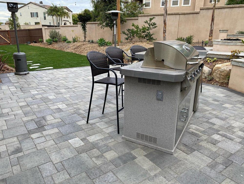 Outdoor patio with a stainless steel grill and black bar stools on a gray paver terrace.