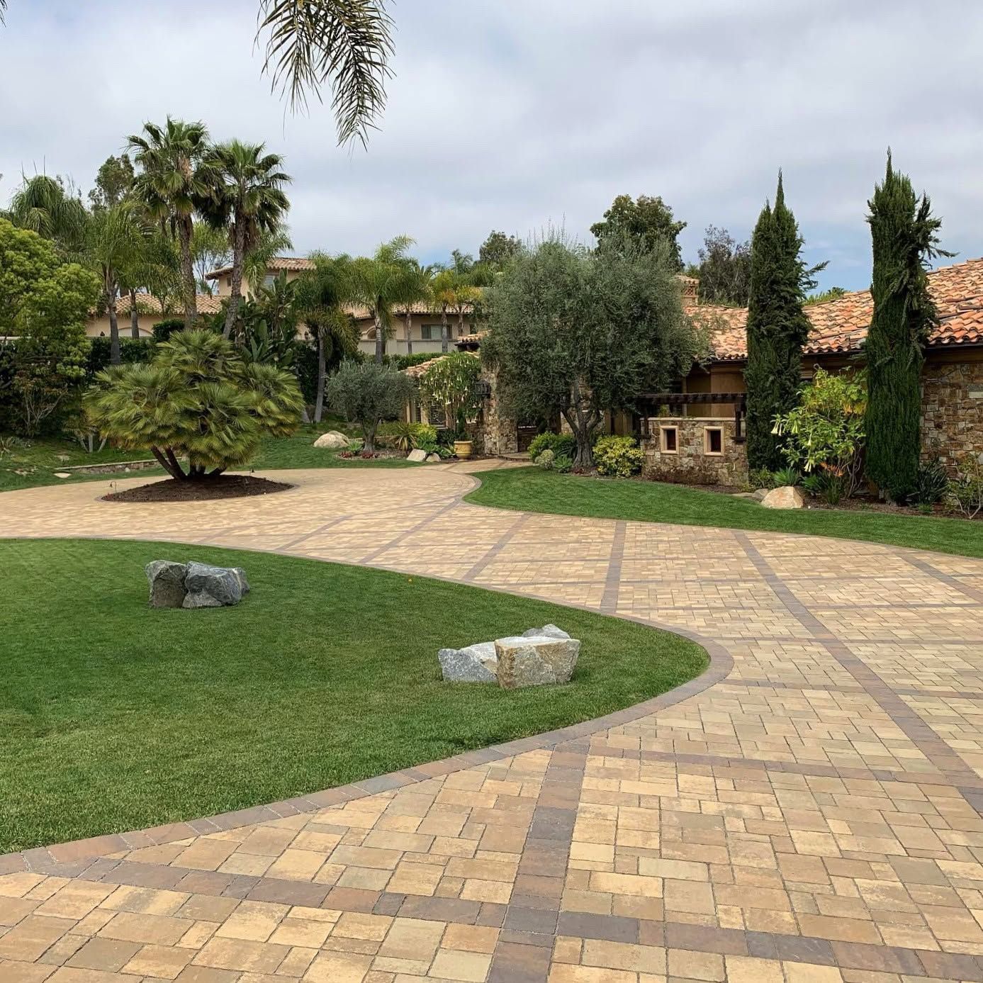 Brick-paved garden path through a green landscaped yard with trees, shrubs, and small stone features