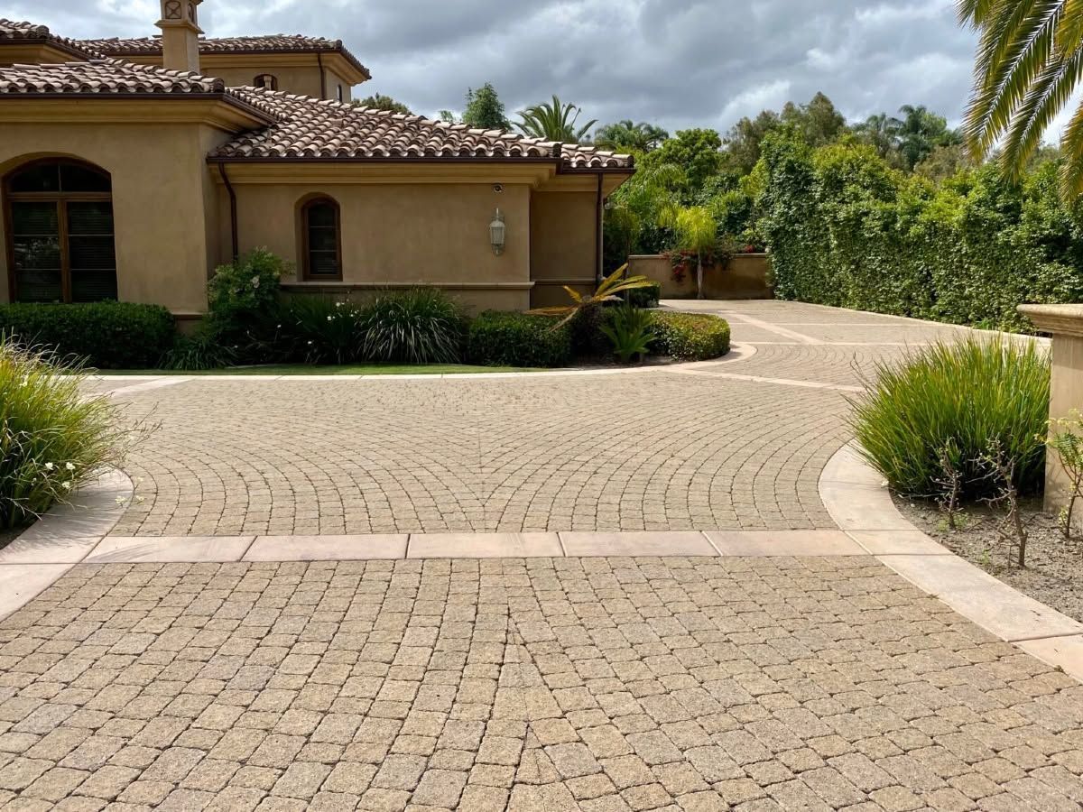 Stone driveway leading to a tan stucco house with a tile roof and desert landscaping under cloudy skies