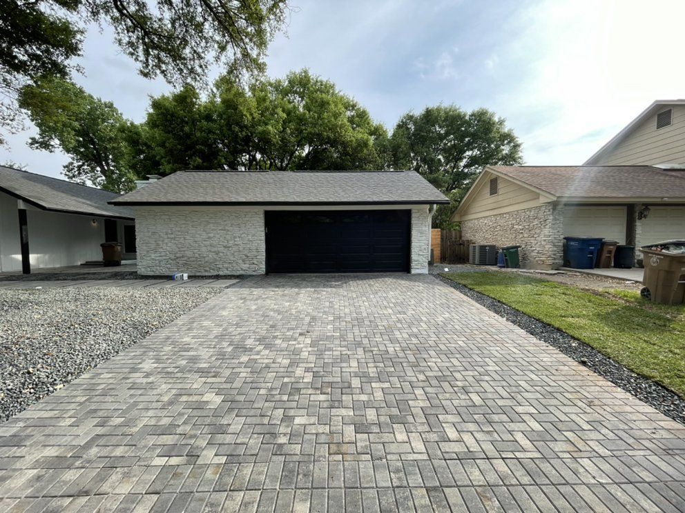 Brick-paved driveway leading to a detached garage with a black door, flanked by neighboring houses and trees
