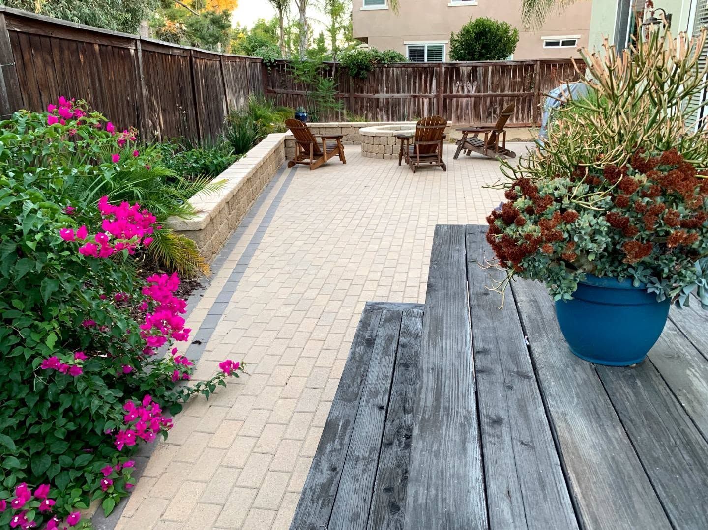 Patio with wooden deck, paver walkway, outdoor chairs, and colorful potted flowers in a fenced yard
